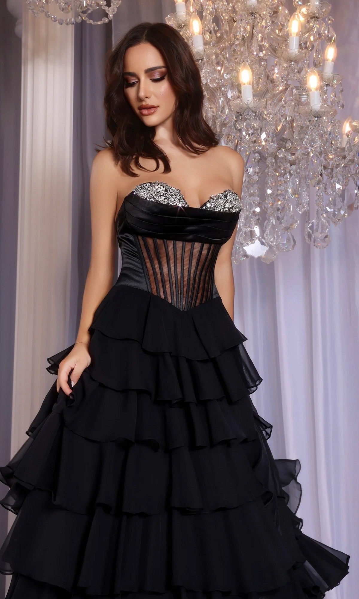 A woman wearing the Portia and Scarlett PS25961 long prom dress with a beaded corset bodice and tiered ruffle skirt stands before crystal chandeliers and white drapery.